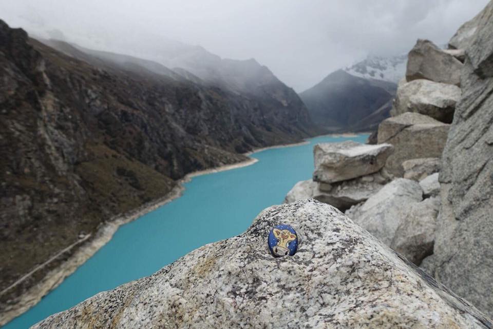 Cholula, a hand-painted cow pebble, on a rock above the turquoise water of Lake Parón in Peru
